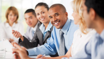 Large group of business people sitting in a row and communicating.  The focus is on African-American looking at the camera.